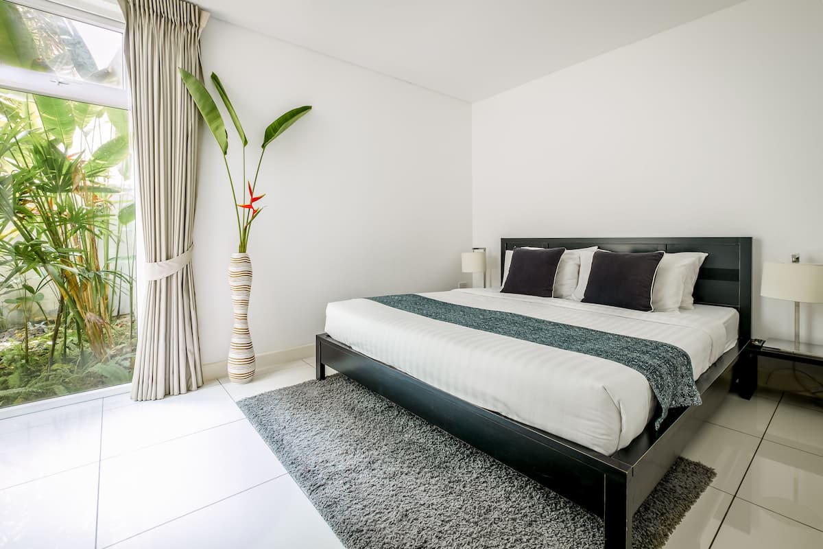 A modern bedroom with a large bed, a bedside table with lamps, a window with curtains, and a potted plant.