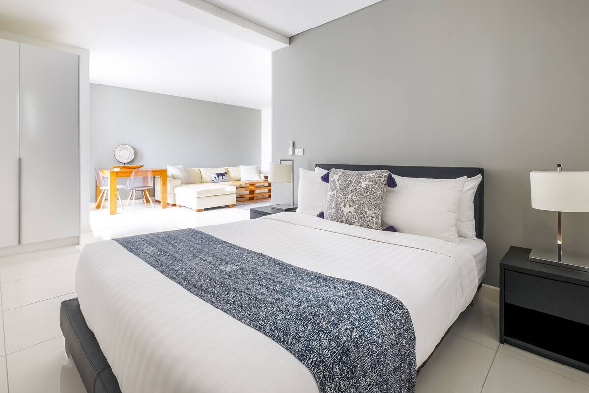 A modern bedroom with a large bed, bedside table, and a desk area with chairs.
