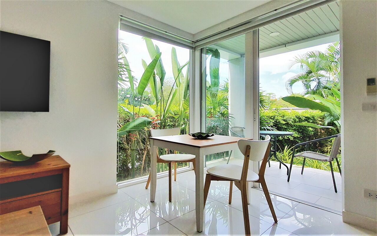 Une chambre moderne dotée d’une table et de chaises, d’un téléviseur fixé au mur et d’une grande porte-fenêtre coulissante donnant sur un balcon agrémenté de plantes tropicales.