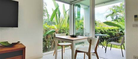 Une chambre moderne dotée d’une table et de chaises, d’un téléviseur fixé au mur et d’une grande porte-fenêtre coulissante donnant sur un balcon agrémenté de plantes tropicales.