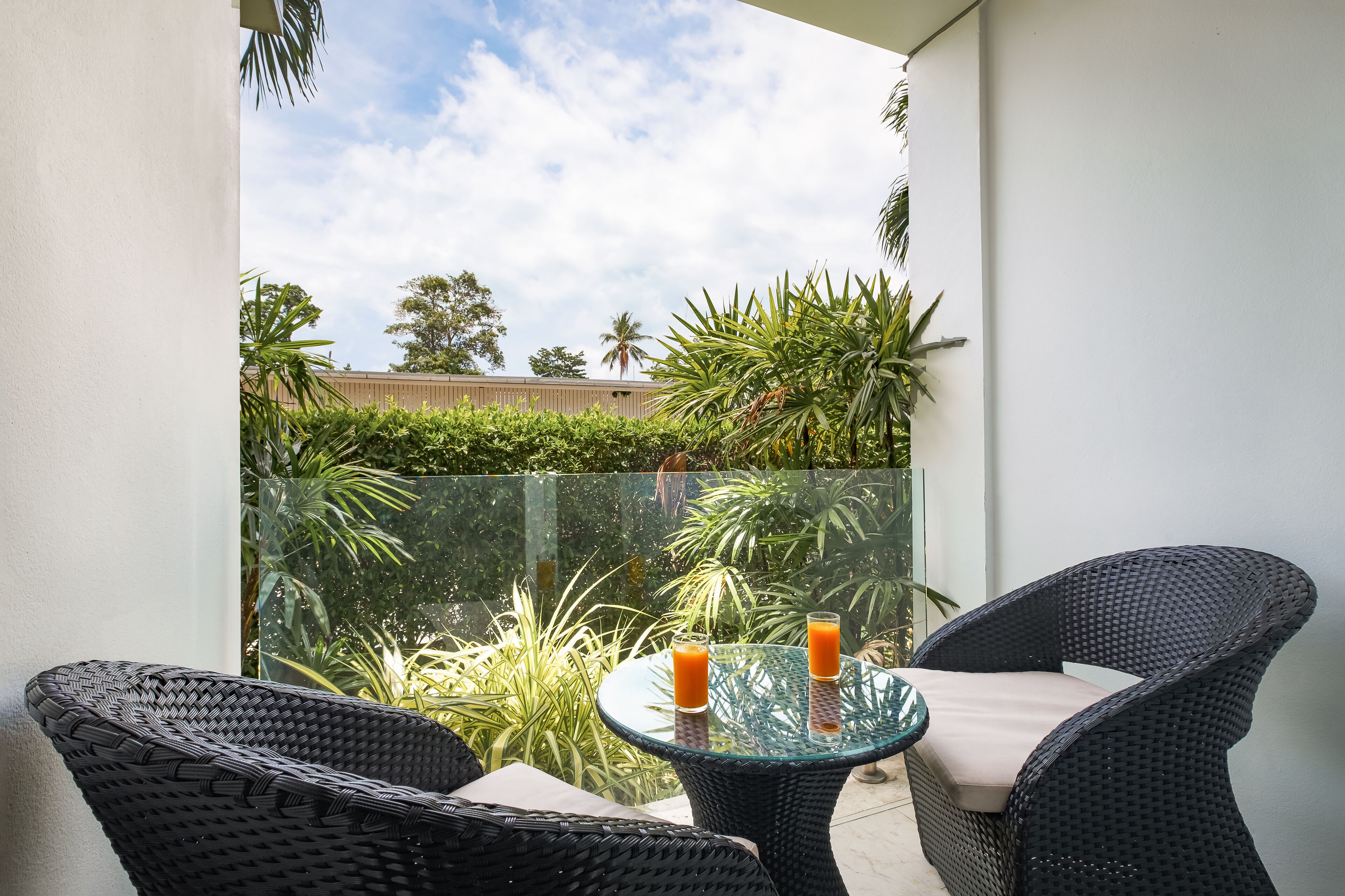 Un balcón con mobiliario de mimbre y una mesa de vidrio, con dos bebidas y vistas a zonas verdes y un edificio al fondo.