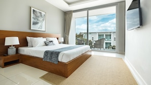 A modern bedroom with a large bed, a bedside table with a lamp, a TV mounted on the wall, and a large window offering a view of the outside. - Horizon Residence Rentals (Koh Samui)