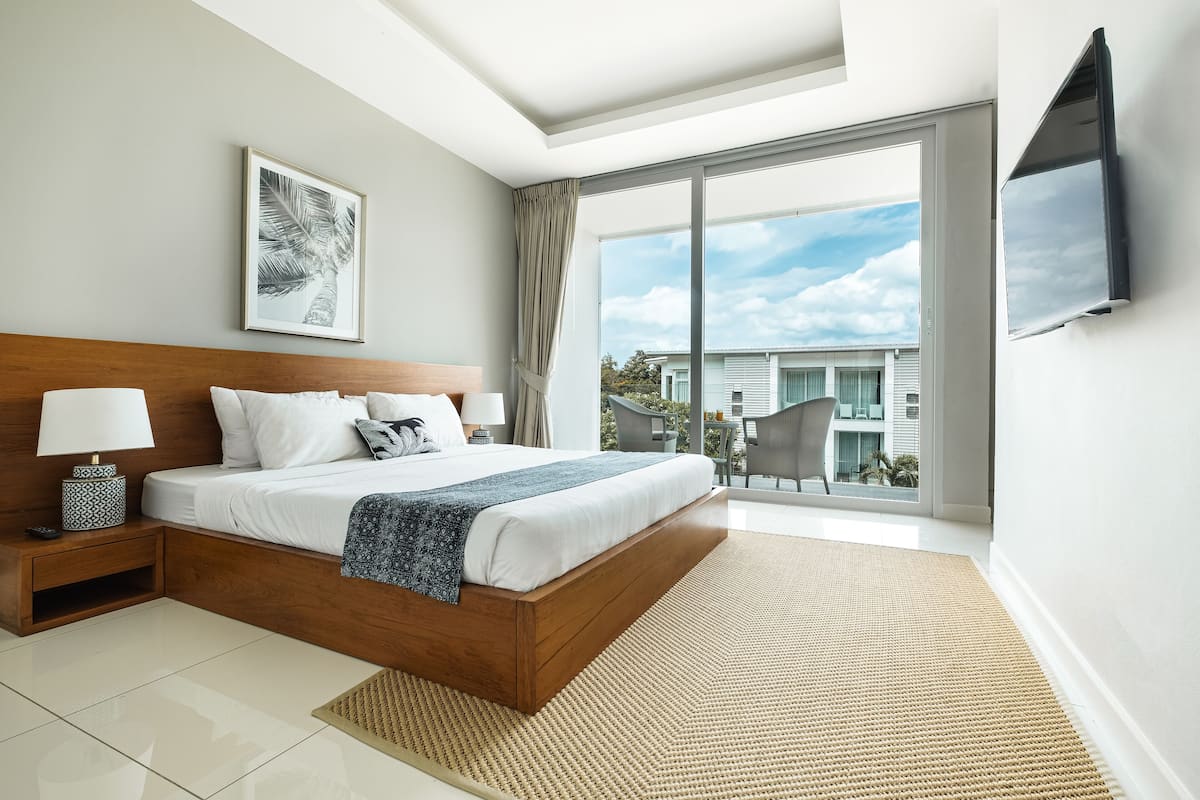 A modern bedroom with a large bed, a bedside table with a lamp, a TV mounted on the wall, and a large window offering a view of the outside.