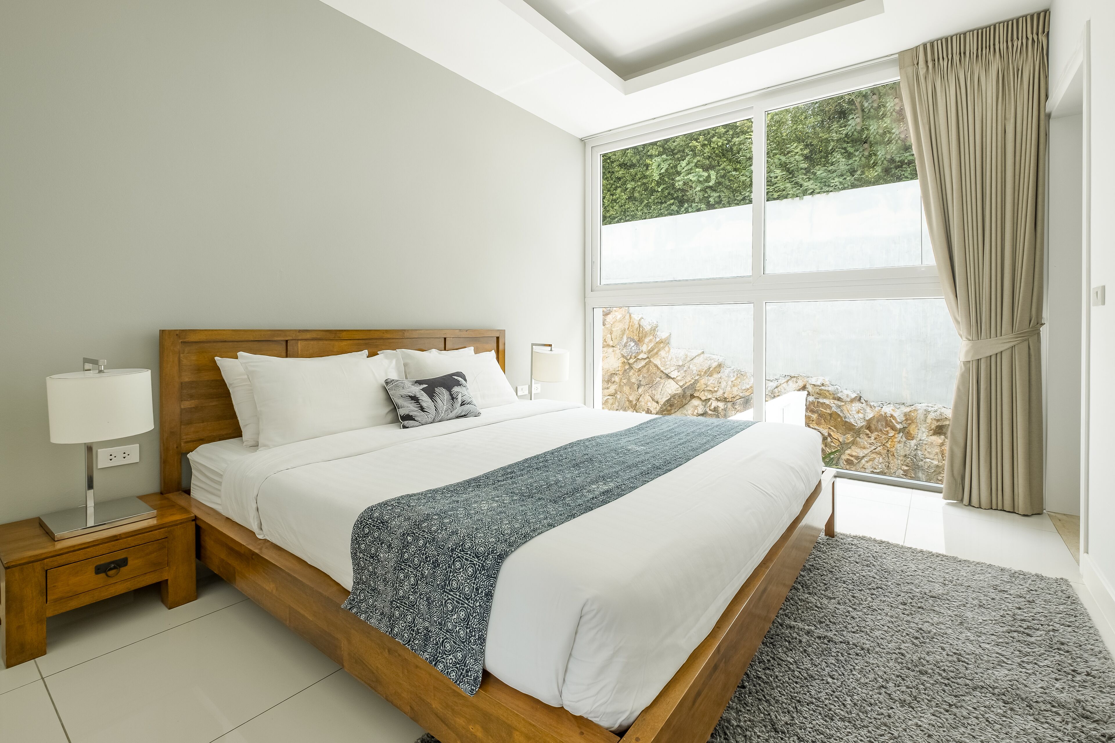 A bedroom with a wooden bed, white bedding, a patterned throw, a nightstand with a lamp, and a large window with curtains.