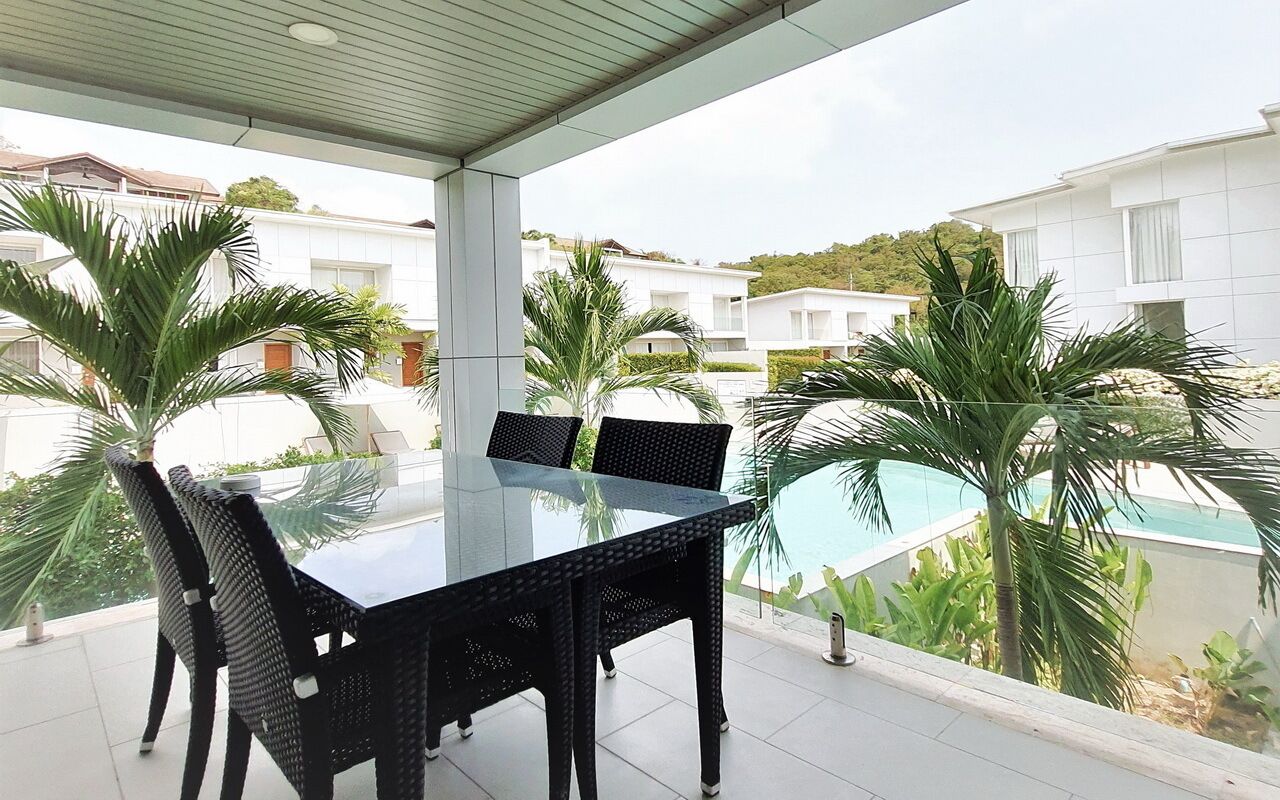 A covered outdoor area with a glass table and black wicker chairs, overlooking a swimming pool and residential buildings.