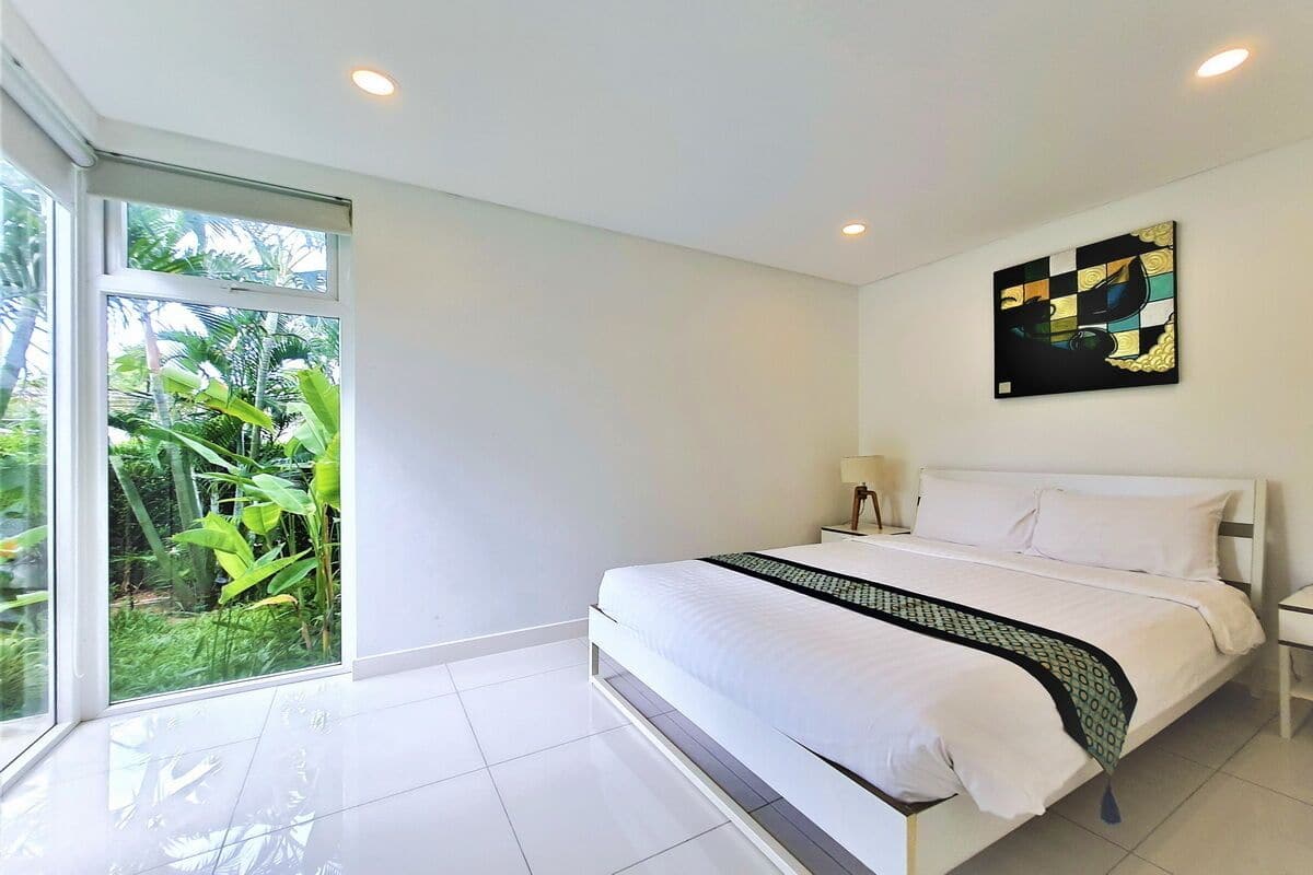 A modern bedroom with a large bed, a window with a view of greenery, and a wall-mounted abstract art piece.