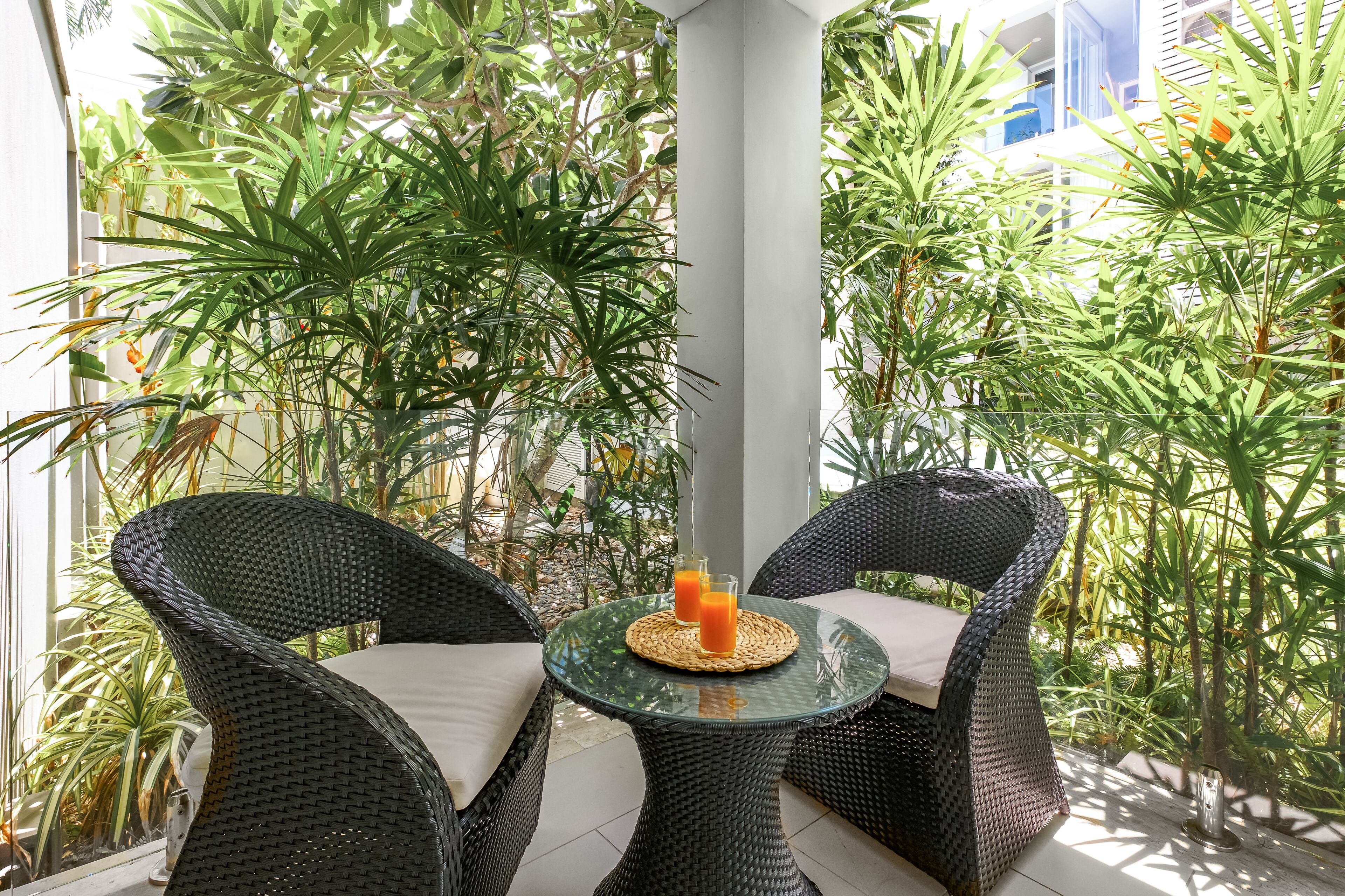 A glass table with wicker chairs and a woven placemat, surrounded by lush tropical plants.