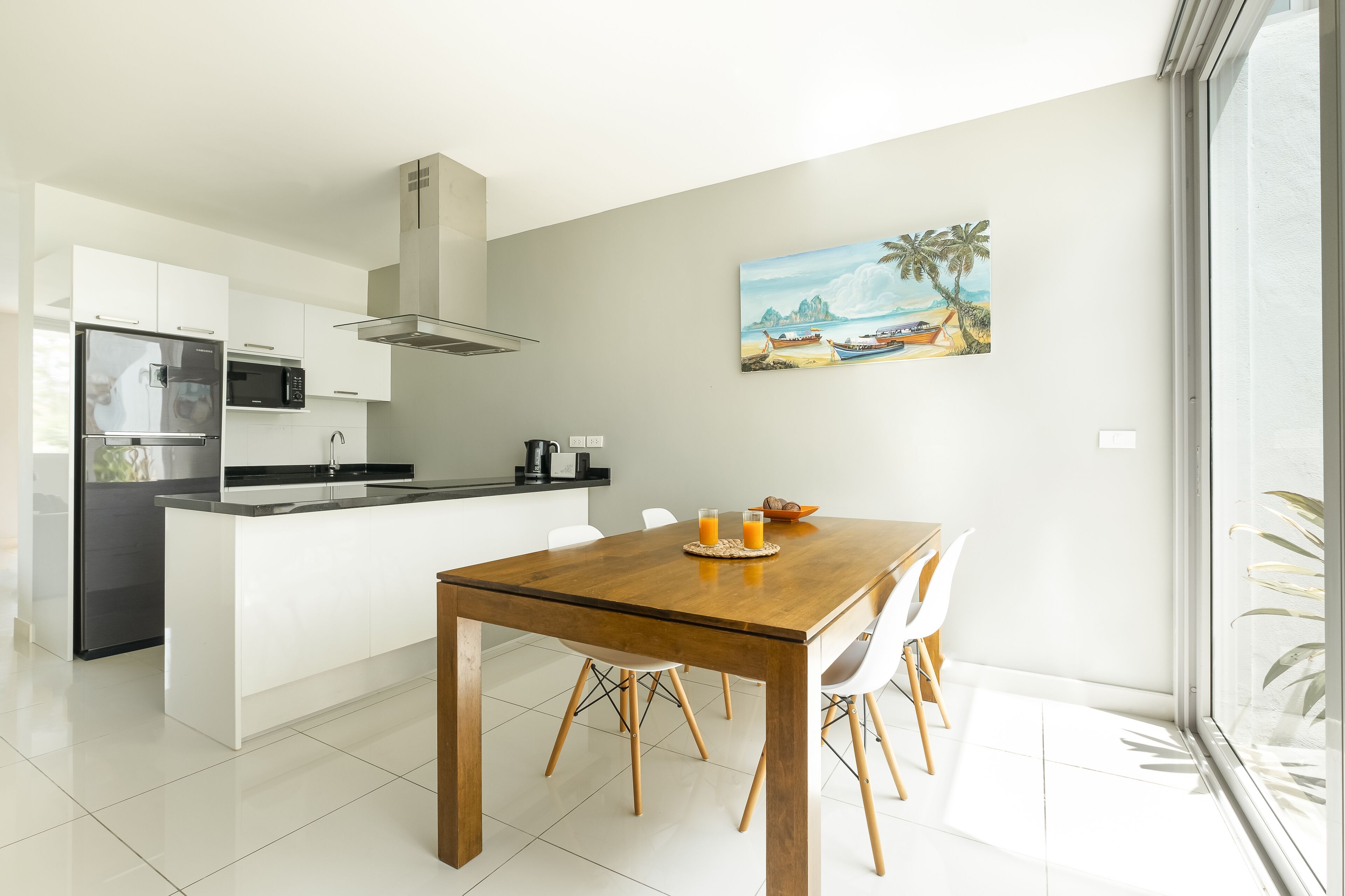 A modern kitchen with white cabinetry, a wooden dining table, and a painting of a beach scene on the wall.