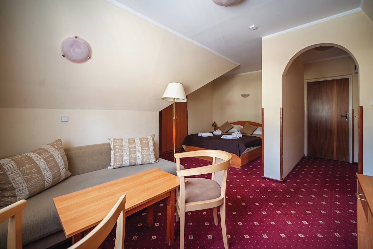Una habitación de hotel pequeña con sofá, una mesita, una silla, cama y un armario de madera.