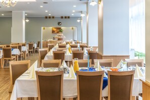 Un comedor amplio con varias mesas dispuestas para el desayuno, con manteles blancos, sillas de madera y diferentes opciones para el desayuno.