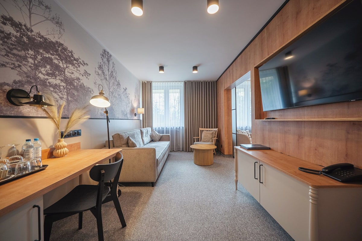 Una habitación de hotel moderna con un escritorio de madera, un sofá, un televisor y un ventanal con cortinas.