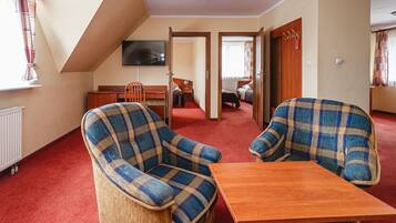 A room with two plaid armchairs, a wooden table, a television on the wall, and a view of another room through an open door.