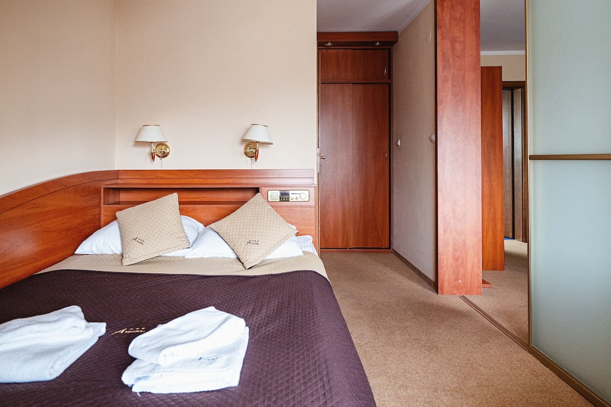 Habitación de hotel con cabecera de madera, dos lámparas fijadas a la pared, cama con colcha oscura, dos almohadas color beige y un par de toallas blancas dobladas sobre la cama.