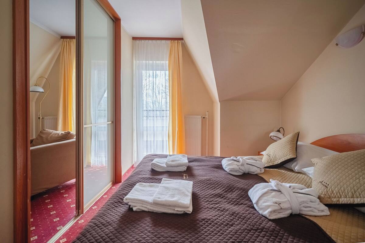 Habitación de hotel con cama, dos almohadas, mesita de noche con lámpara, ventana con cortinas y radiador.