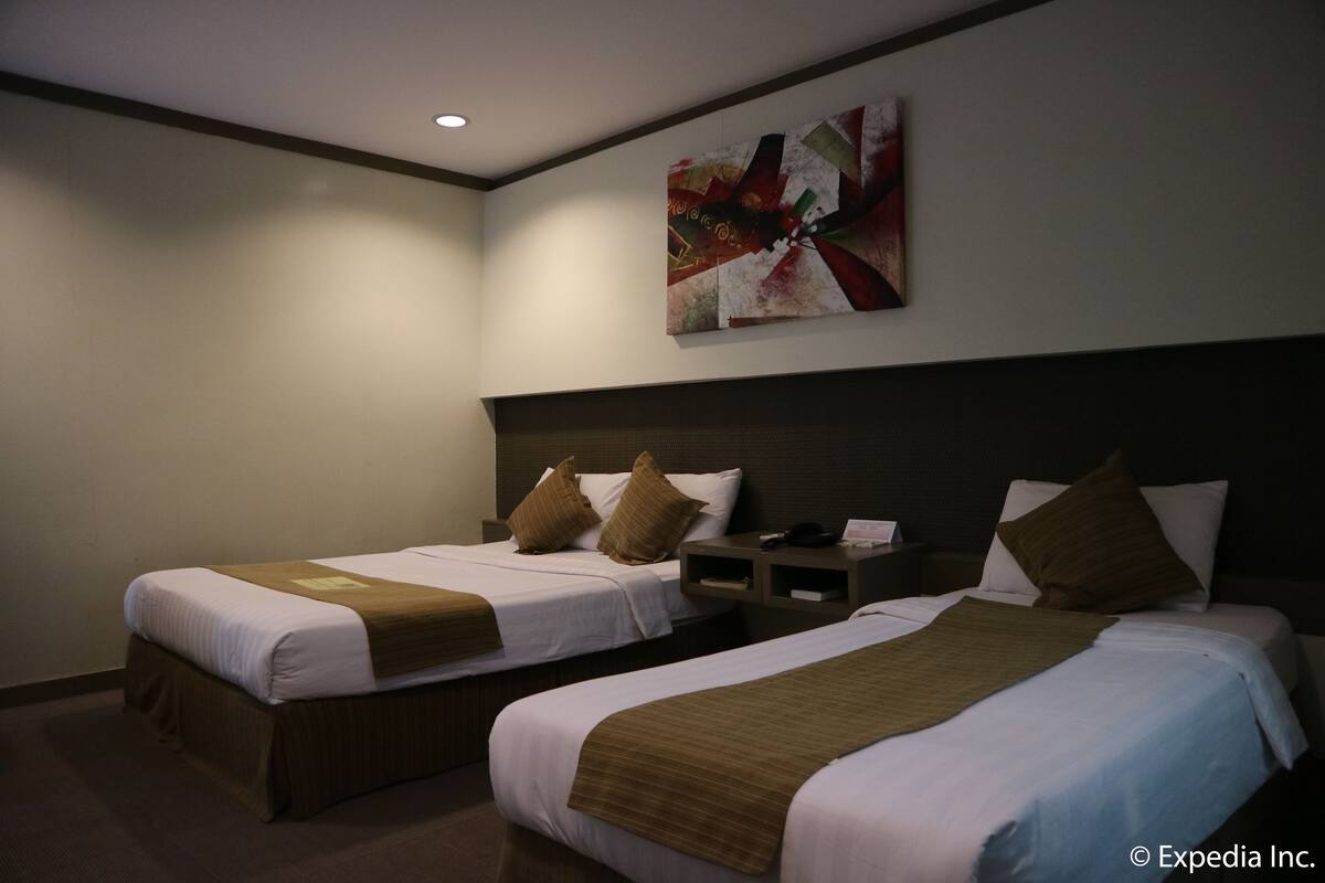 A hotel room with two beds, each with a brown headboard and white bedding. There is a painting on the wall above the beds.