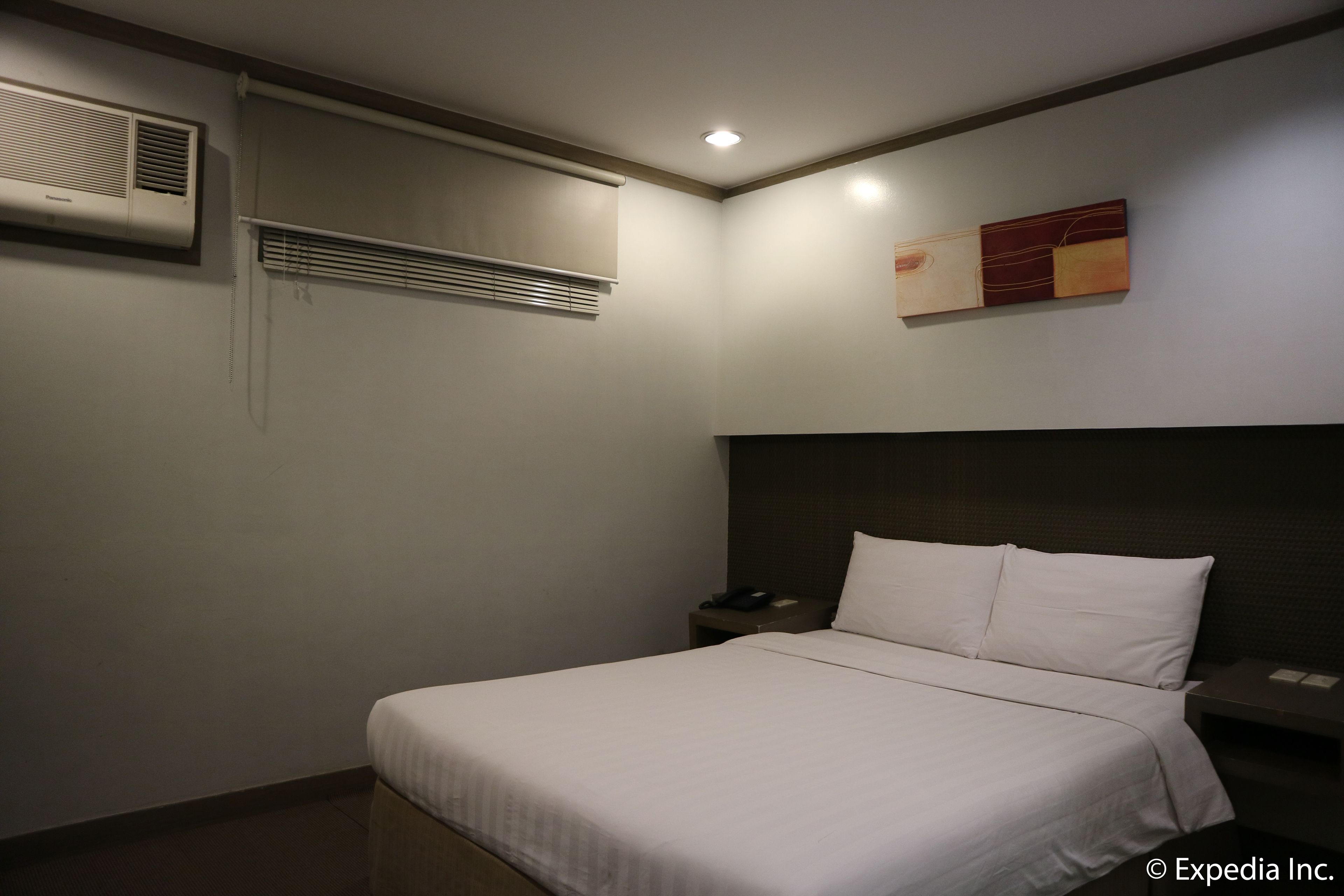 A hotel room with a bed, bedside tables, a wall-mounted air conditioner, a window with blinds, and a painting on the wall.
