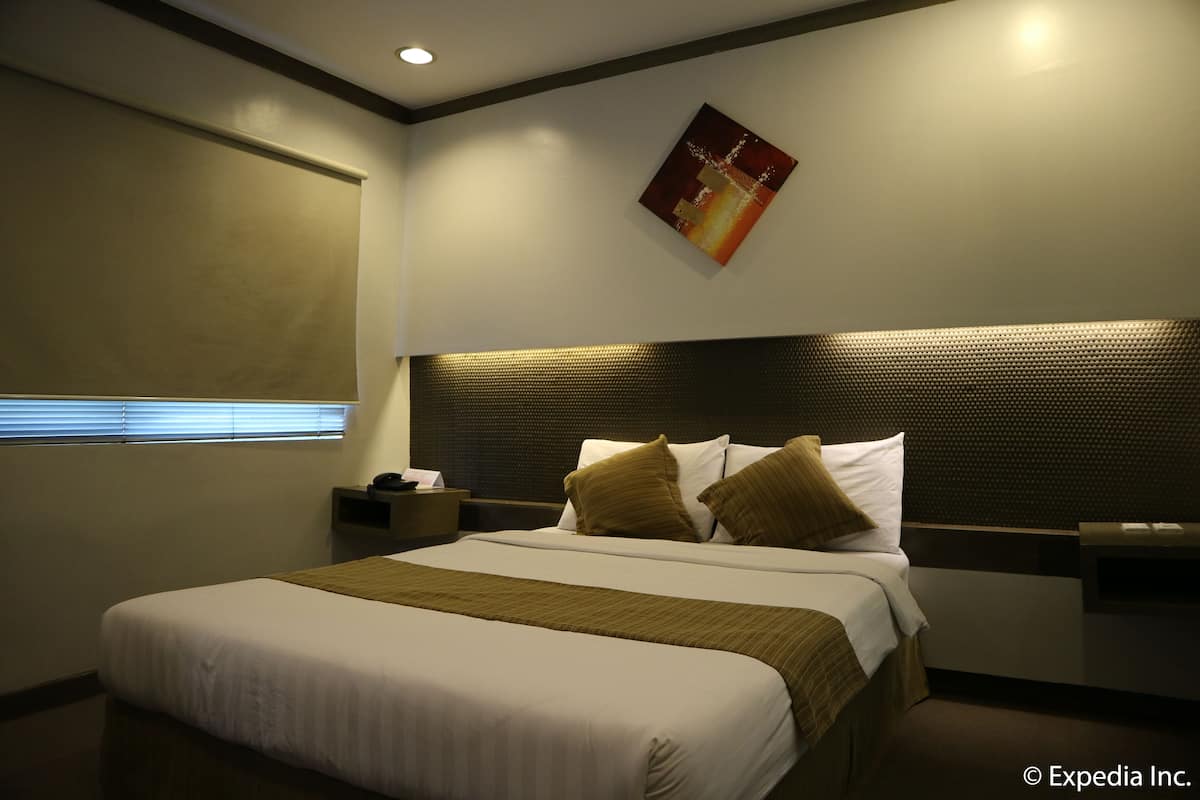 A hotel room with a large bed, a headboard, a nightstand, a bedside table, and a window with blinds.