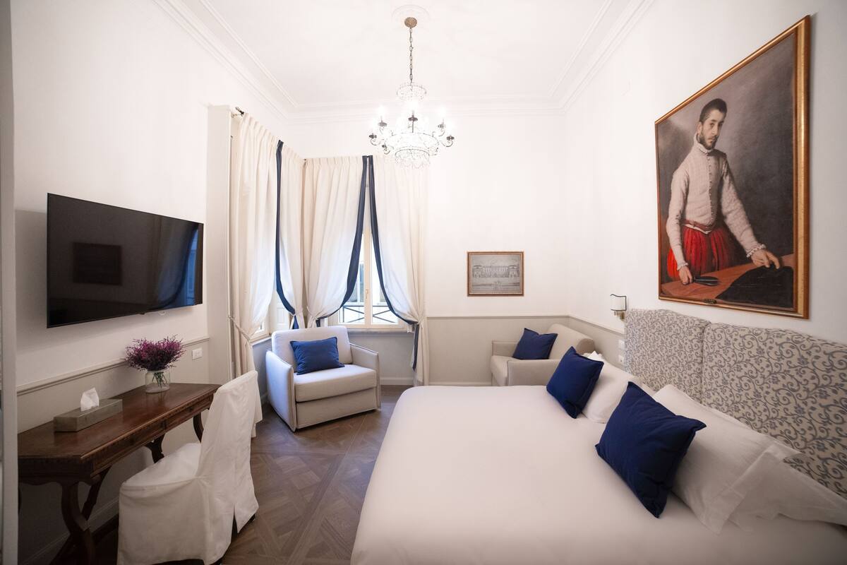 A hotel room with a large bed, a TV, a desk with a vase of flowers, and a painting on the wall.