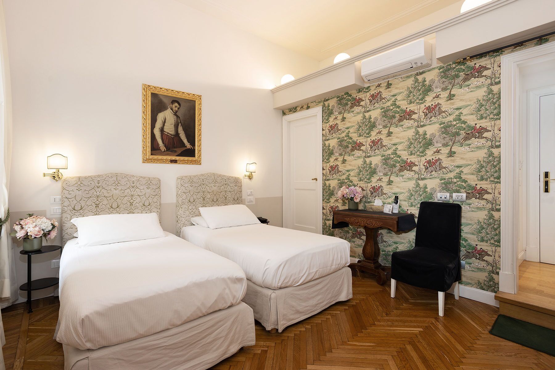 A hotel room with two beds, a portrait on the wall, a floral wallpaper, and a wooden floor.
