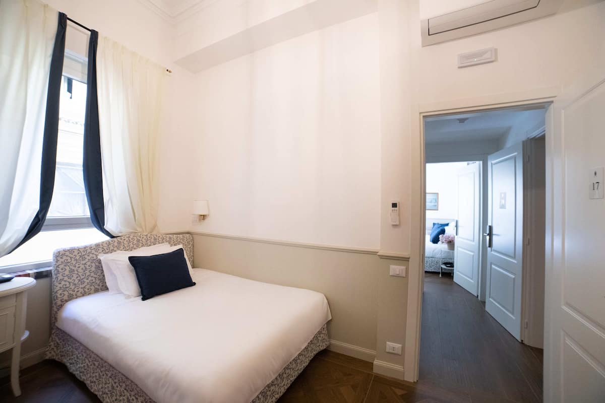 A bedroom with a bed, a nightstand, a window with curtains, and a door leading to another room.