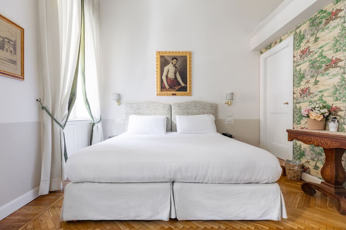 A neatly made bed with white linens, a headboard with a decorative panel, a framed portrait on the wall, and a wooden nightstand with a vase of flowers.