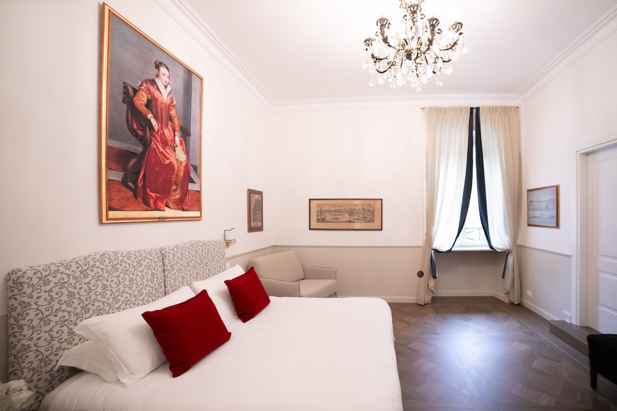 Una habitación de hotel con una cama grande, una araña de cristal, un cuadro en la pared y un ventanal con cortinas.