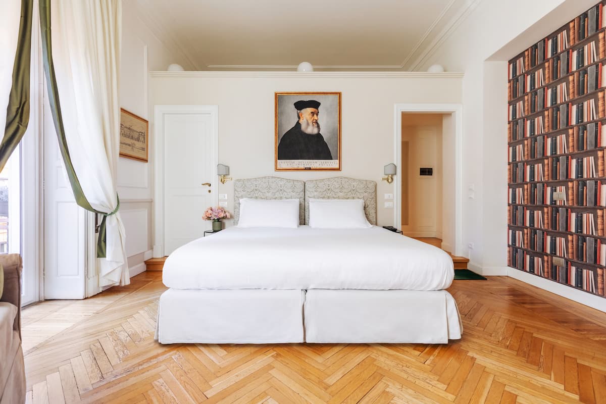 A large bed with a headboard, a wooden herringbone floor, a portrait above the bed, and a bookshelf to the right.