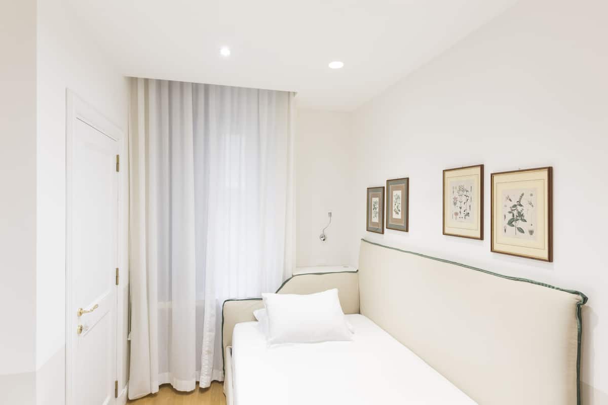 A bedroom with a bed, a door, and a window with curtains. There are framed pictures on the wall.
