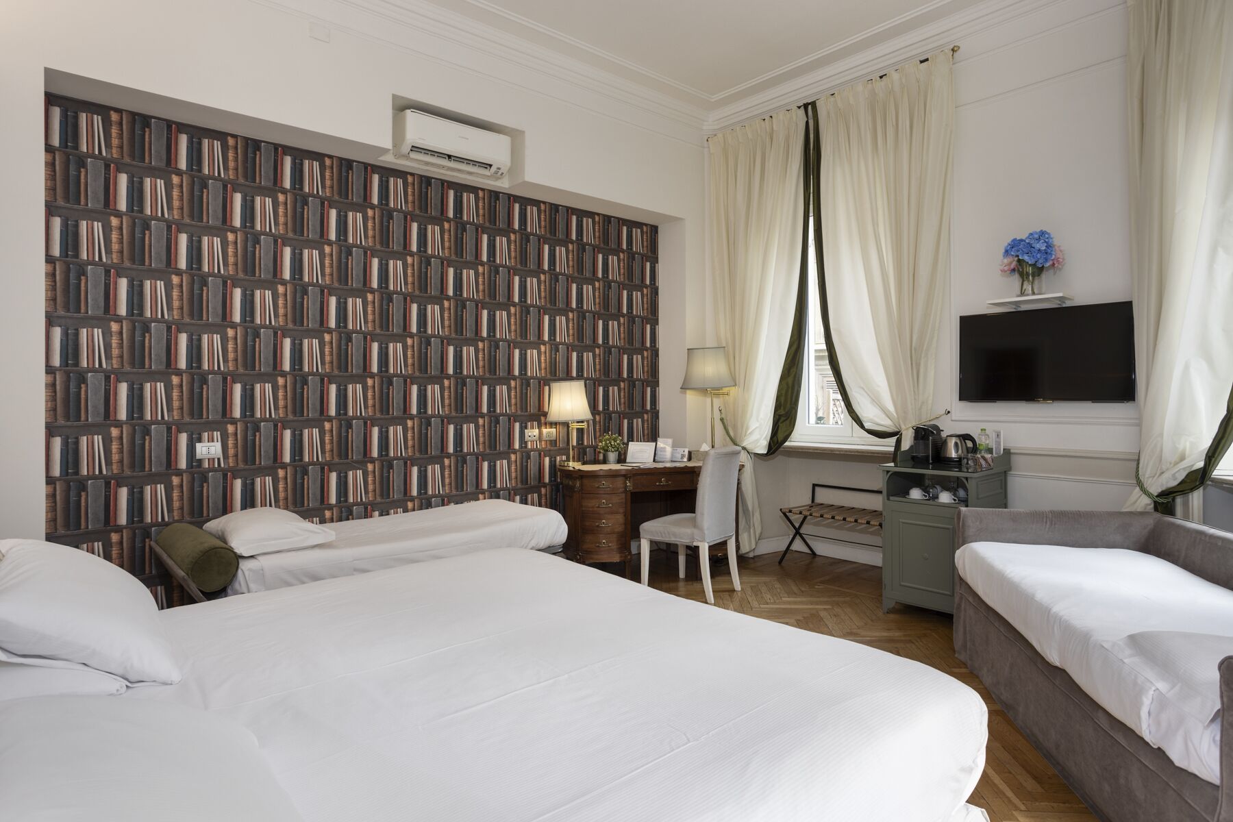 Habitación de hotel con dos camas, un escritorio con silla, una estantería, un televisor y una ventana con cortinas.