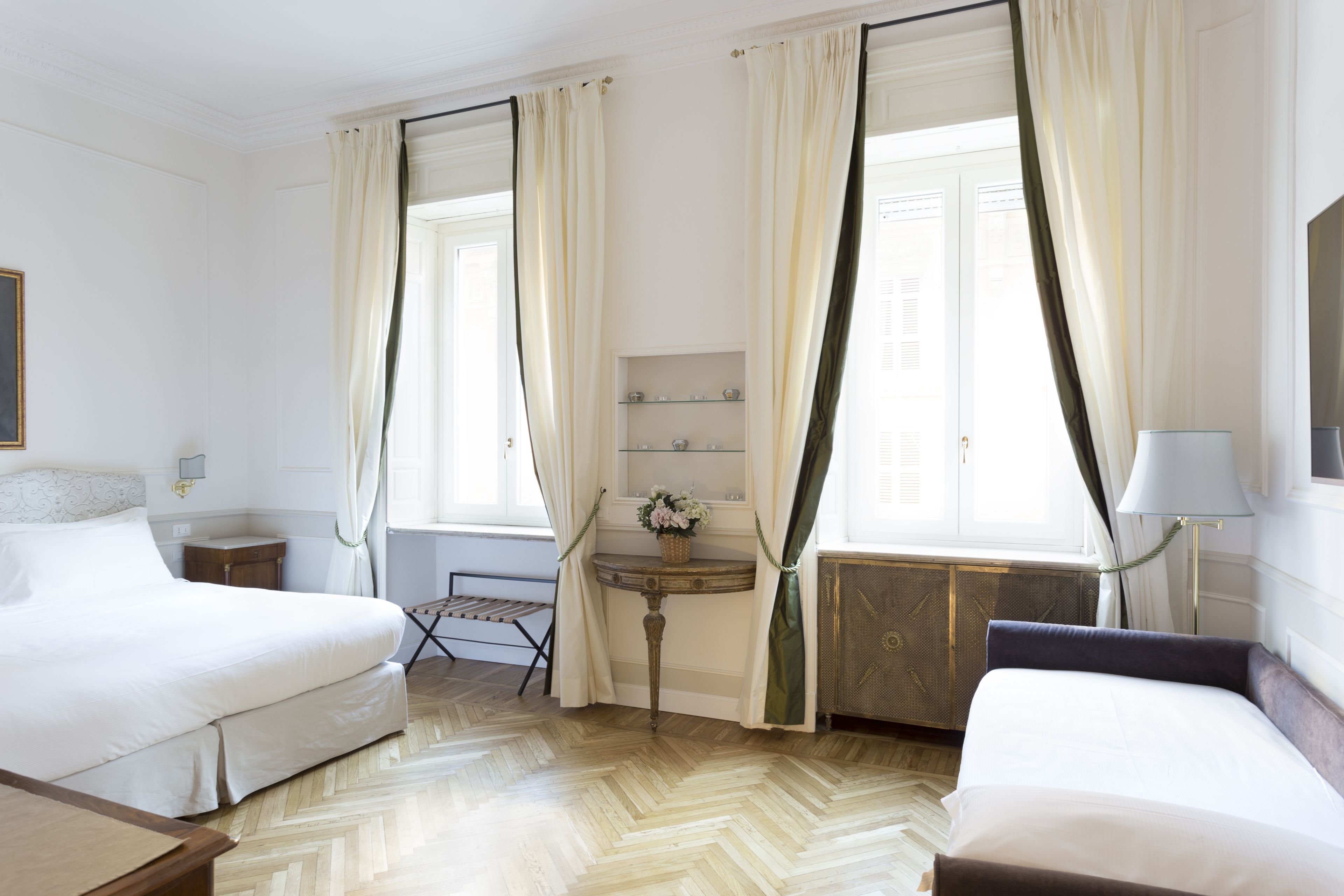 A hotel room with two beds, a small table with a vase of flowers, a lamp, and a radiator.