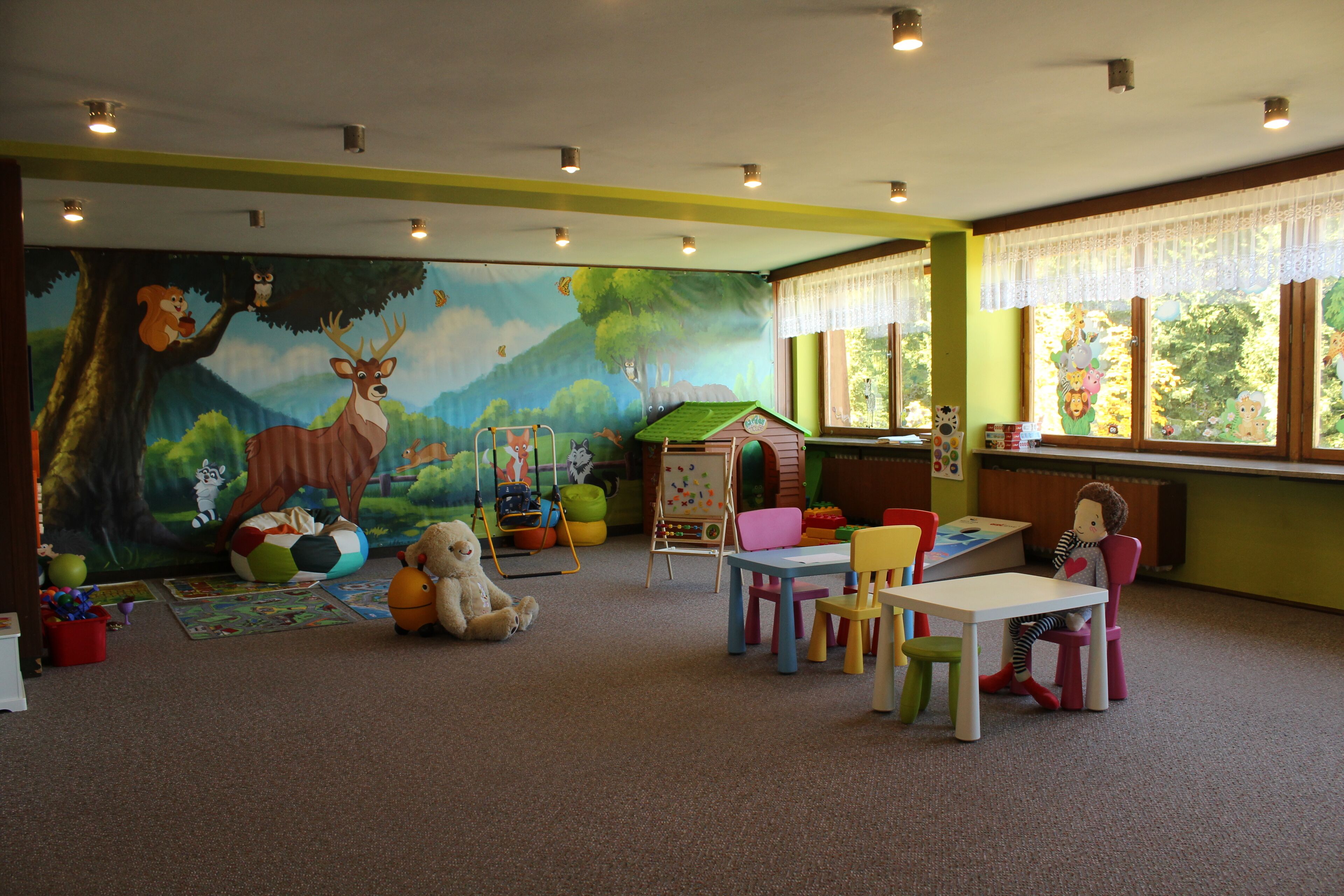 Une salle de jeux pour enfants avec une fresque murale, des meubles colorés et une fenêtre donnant sur des arbres.