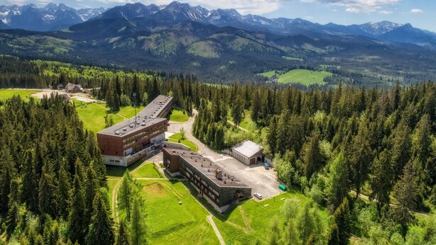 Un bâtiment moderne doté d’un toit plat, entouré d’une forêt dense et avec des montagnes en arrière-plan.