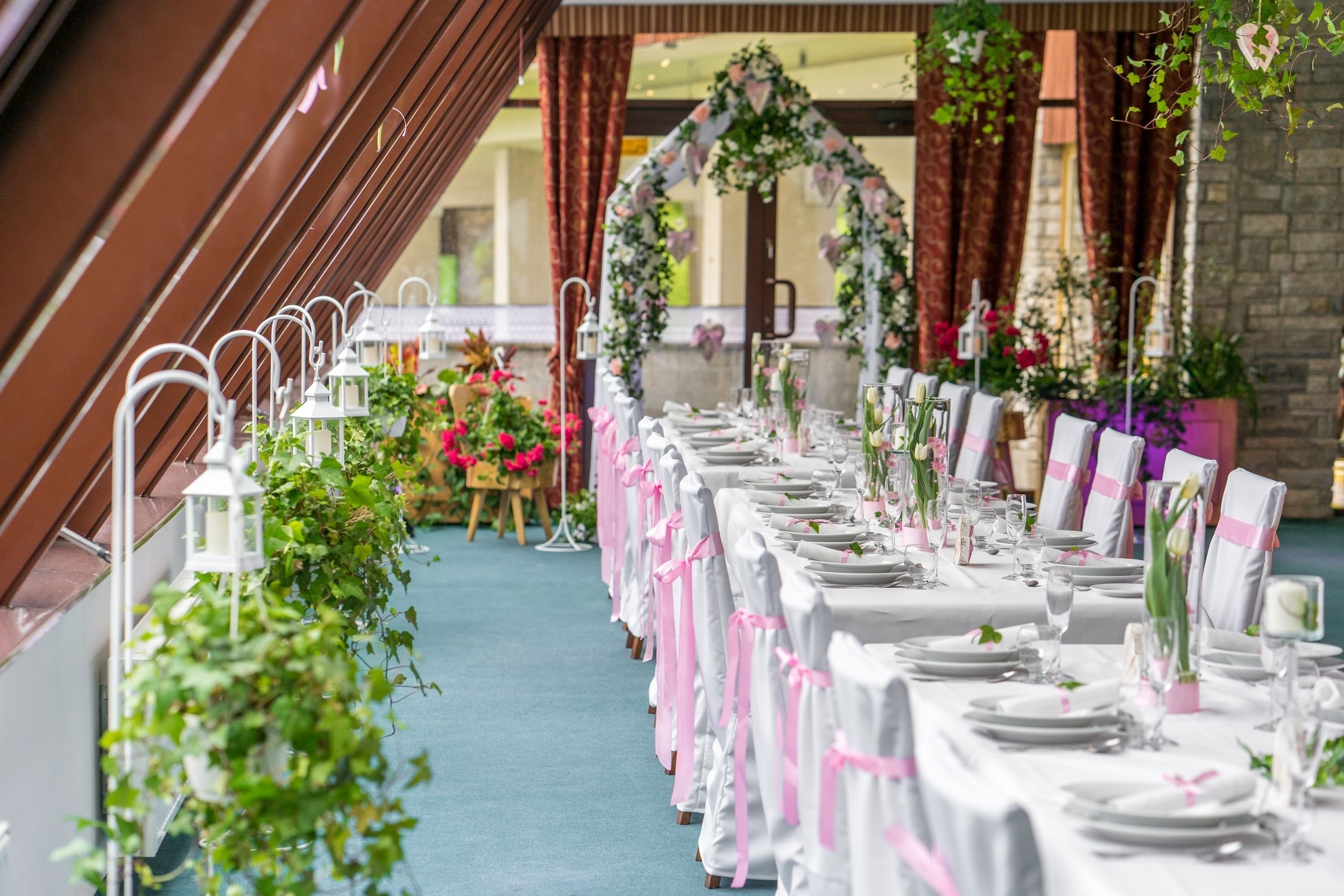 Une longue table de banquet, recouverte de nappes blanches et ornée de rubans roses, est installée dans une pièce dont un mur est en pierre et qui est agrémentée d’une arche fleurie.