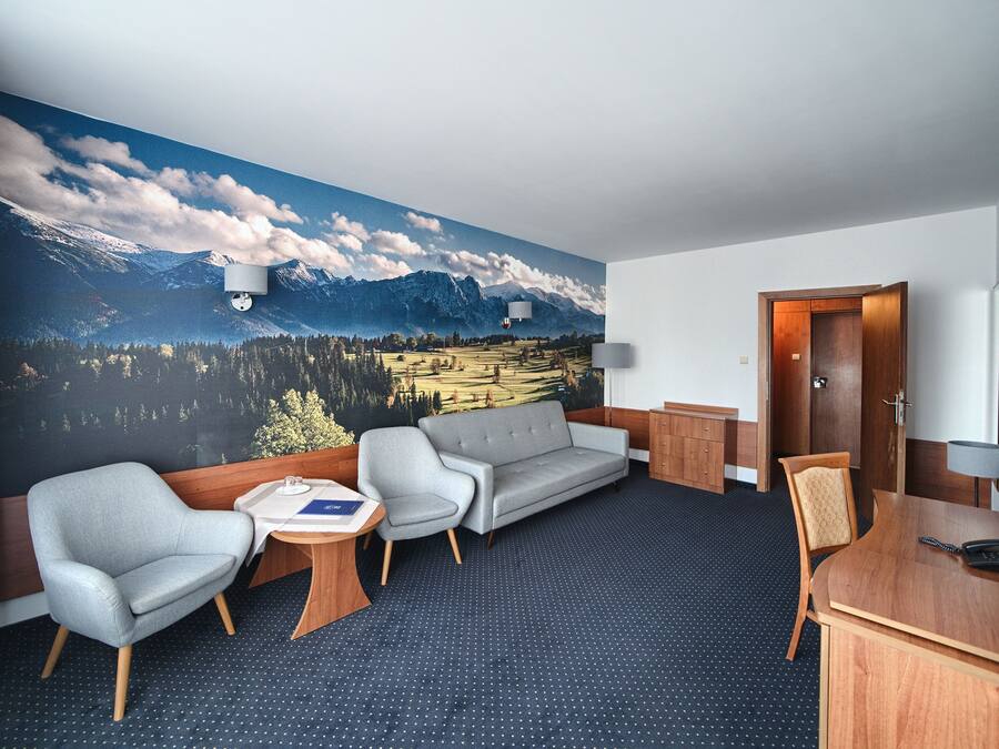 A hotel room with a mountain landscape mural, a grey sofa, two armchairs, a small table, a desk, and a wooden cabinet.