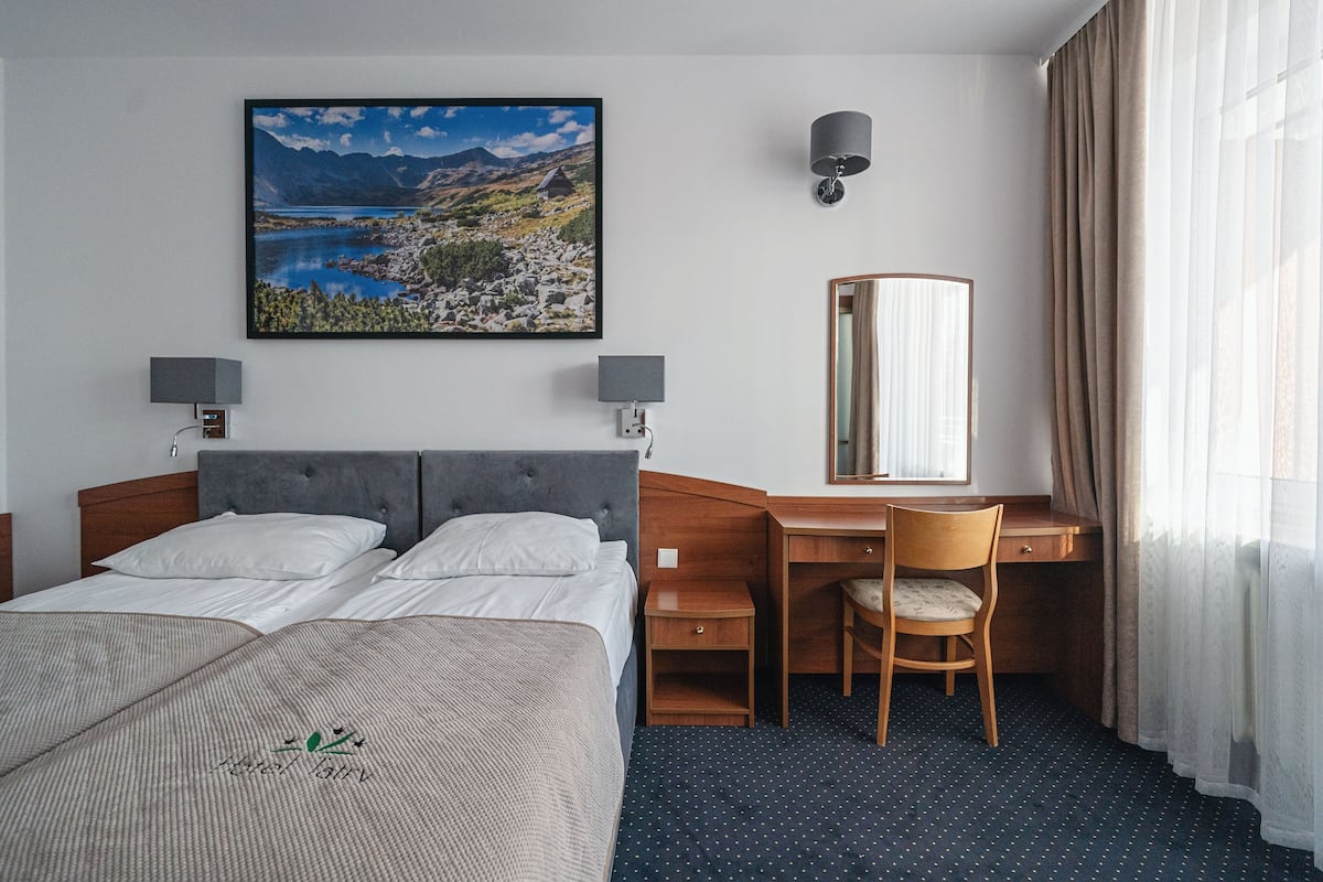 Een hotelkamer met een bed, twee nachtkastjes, een bureau met een stoel, een spiegel en een groot landschap op de muur.