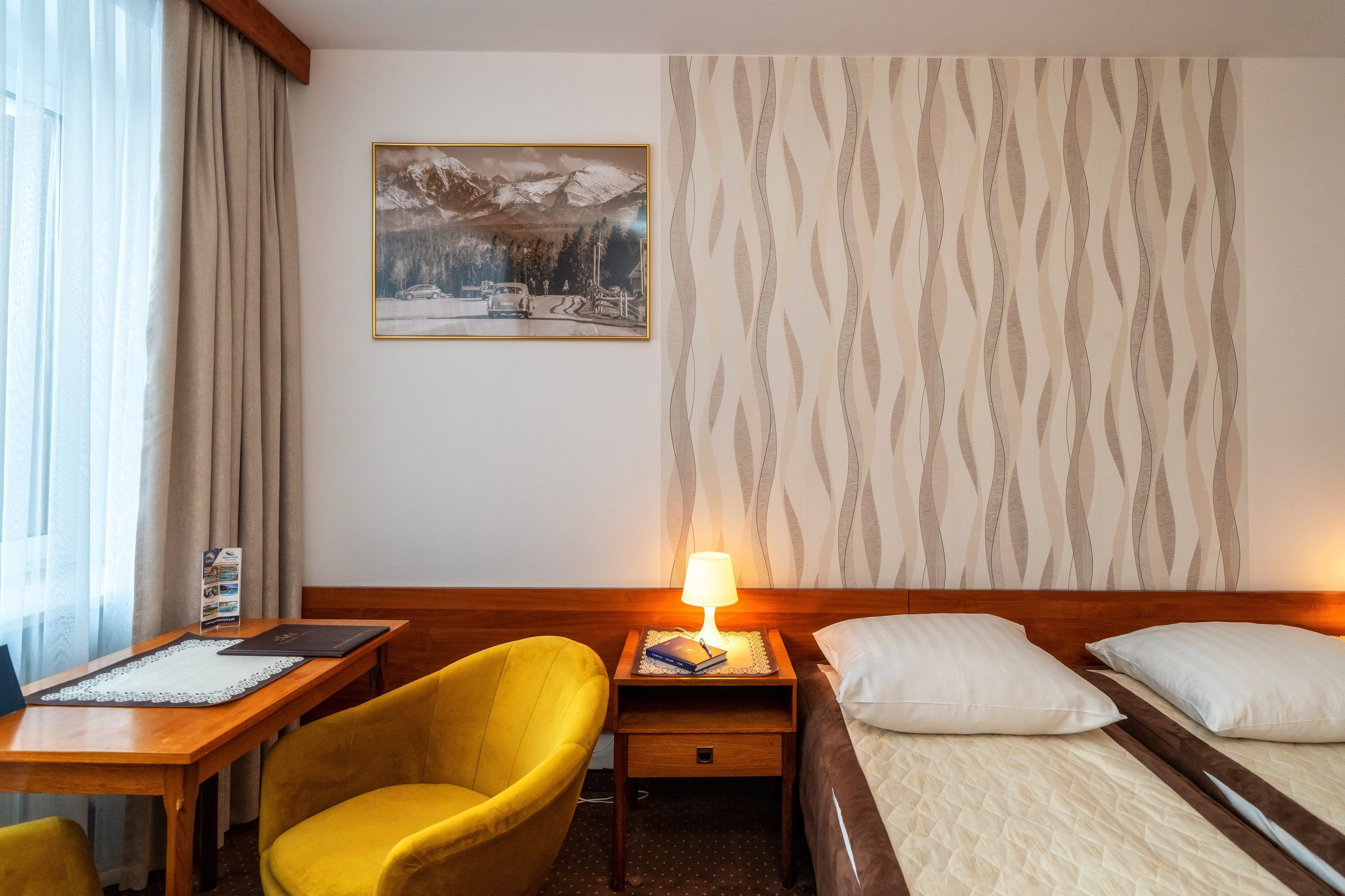 Habitación de hotel con mesita de noche de madera, lámpara, cama con almohadas blancas, sillón amarillo, cuadro en la pared y ventana con cortinas.