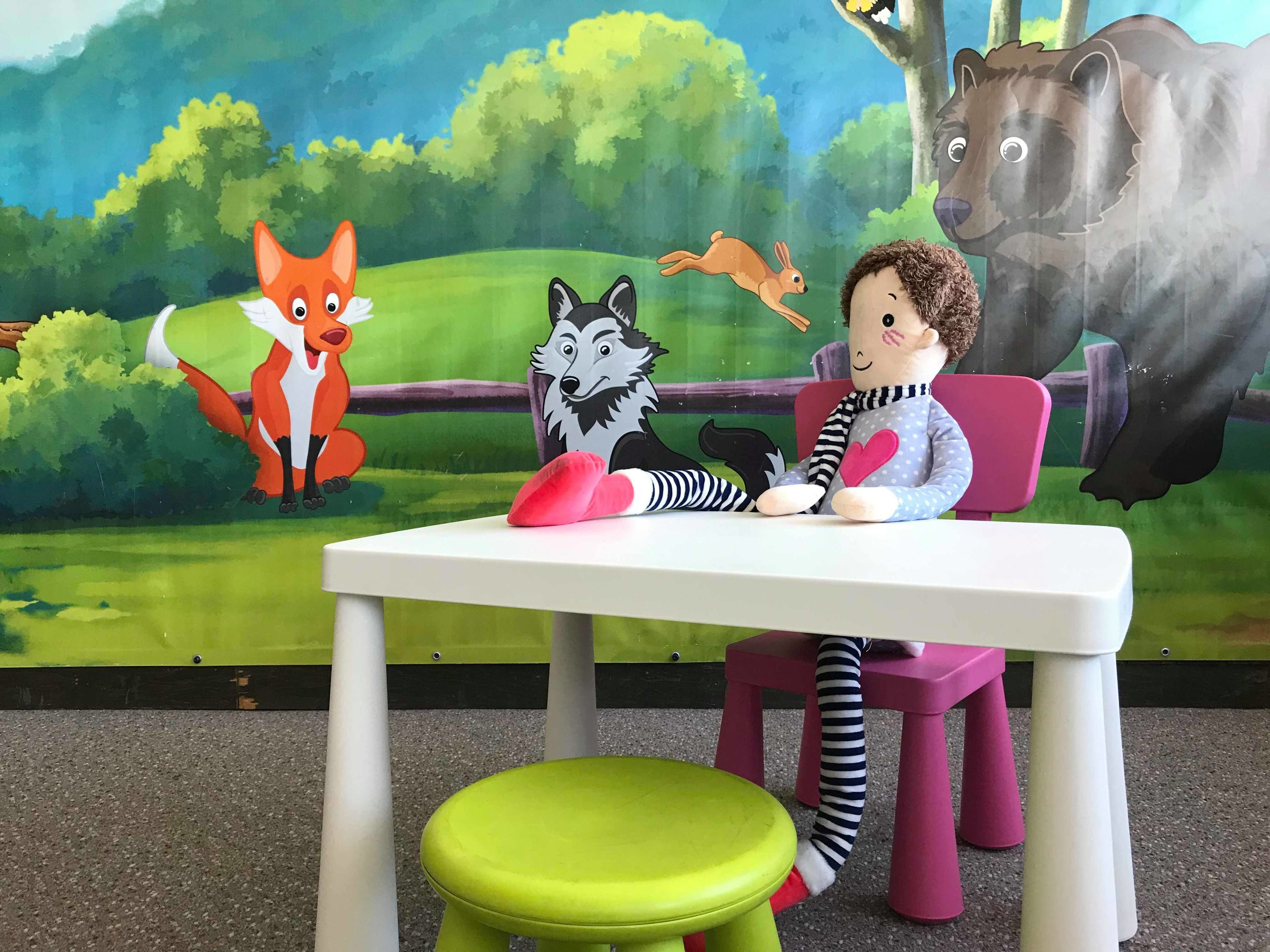 Un enfant est assis à une table, avec un jouet en peluche, un tabouret vert, et une fresque représentant des animaux de la forêt en arrière-plan.