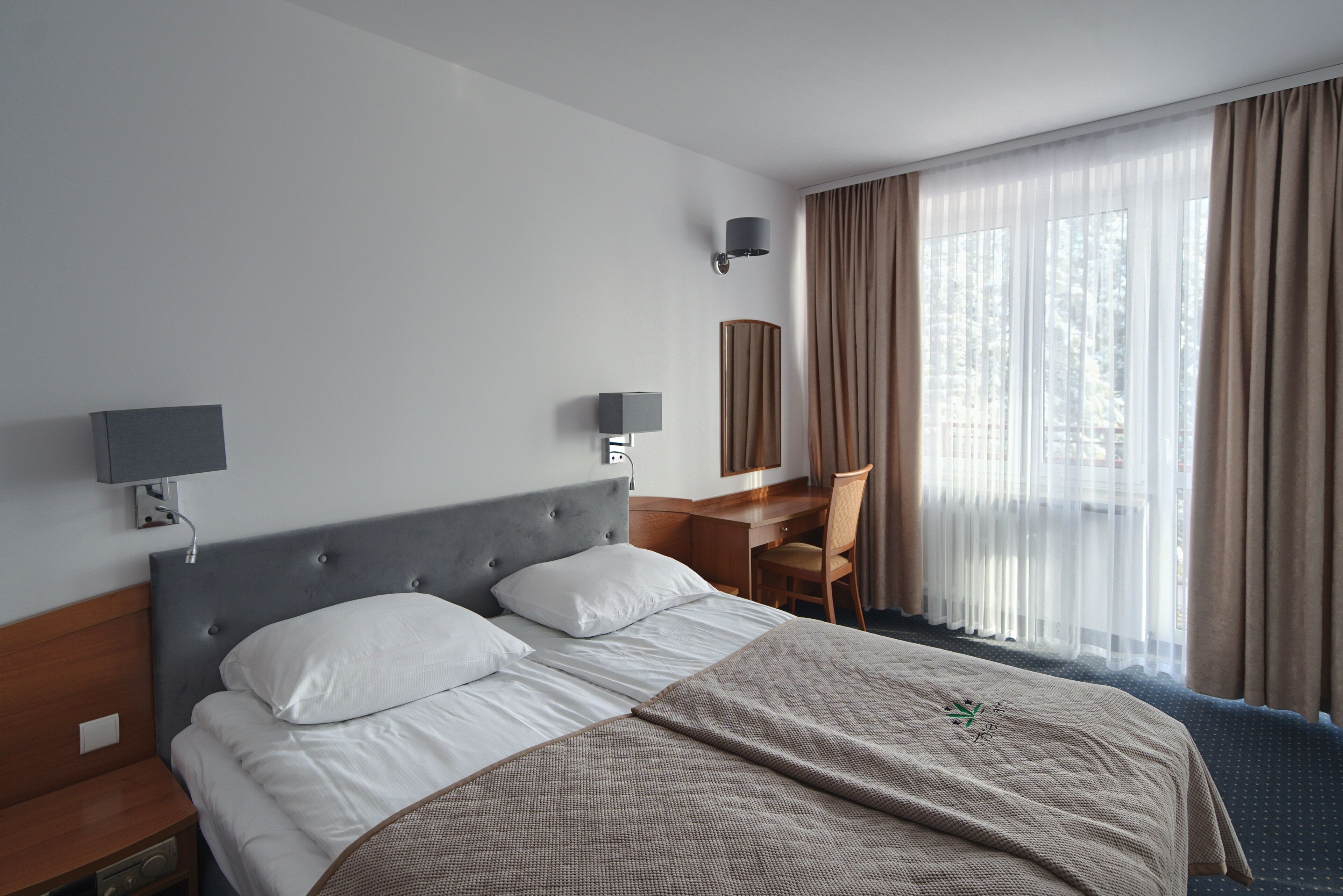 A hotel room with a bed, bedside tables, a desk, a chair, a lamp, and a large window with curtains.
