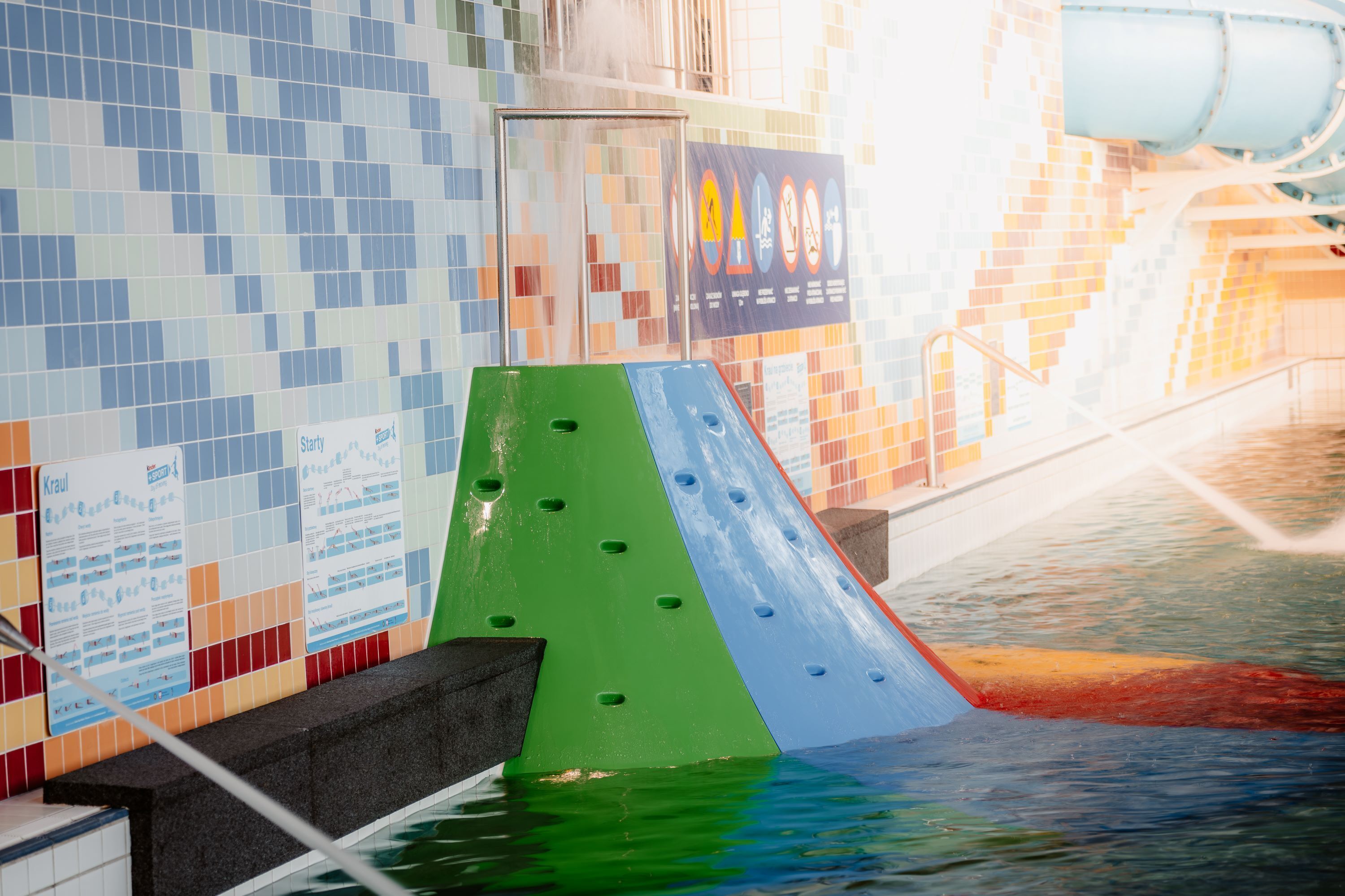 Un toboggan aquatique avec une partie verte et une partie bleue, entouré de murs recouverts de carreaux colorés et agrémenté d’un élément décoratif aquatique.