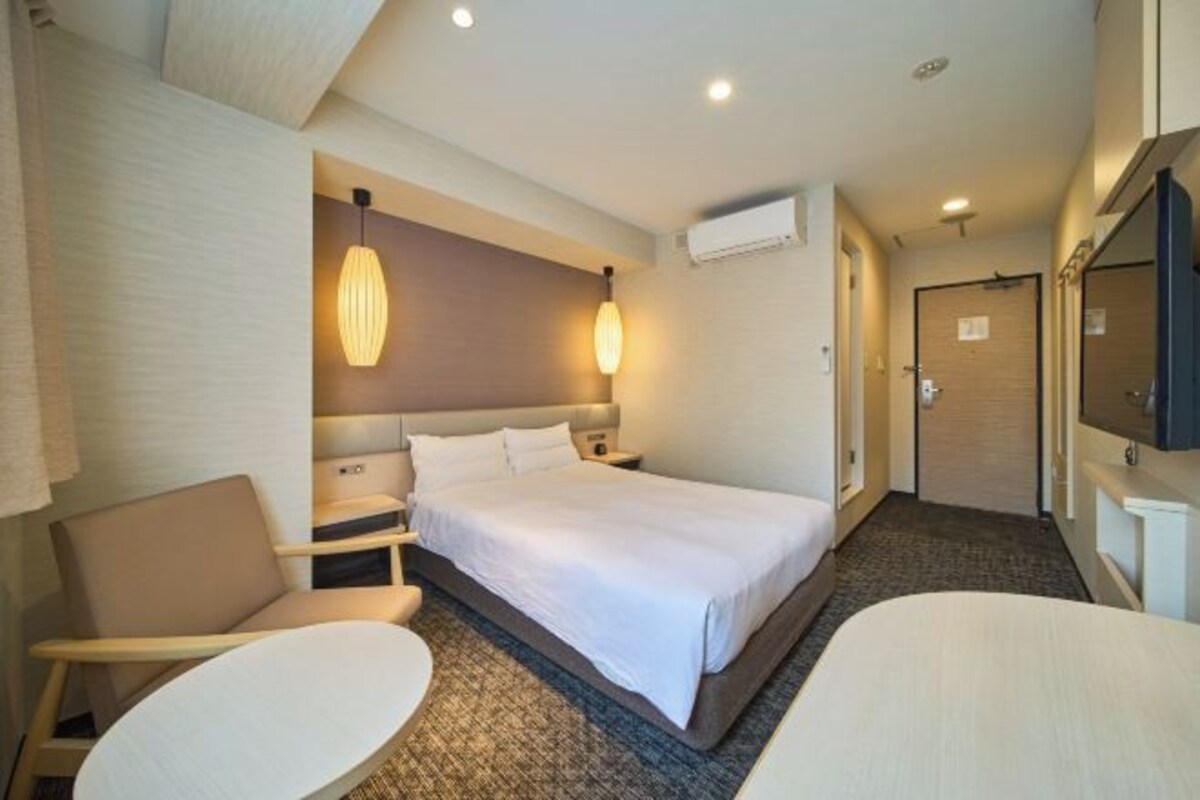 A hotel room with a bed, bedside tables, a chair, a small table, and a TV.