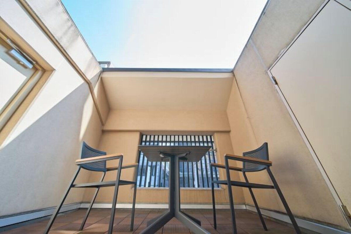 Un pequeño balcón con una mesa y dos sillas, una ventana con rejas y vistas al cielo.