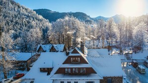 Un edificio cubierto de nieve con techo inclinado, rodeado de un área boscosa y montañas al fondo.