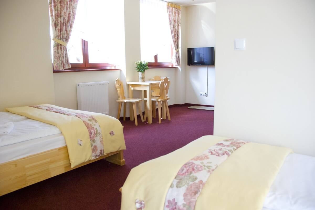 A hotel room with two beds, a dining table with chairs, a TV, and a window with curtains.