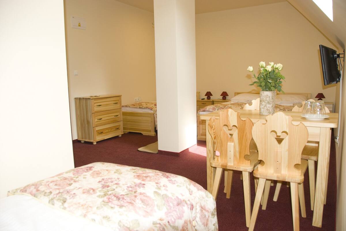A bedroom with a bed, a wooden dresser, a desk with chairs, and a TV mounted on the wall.