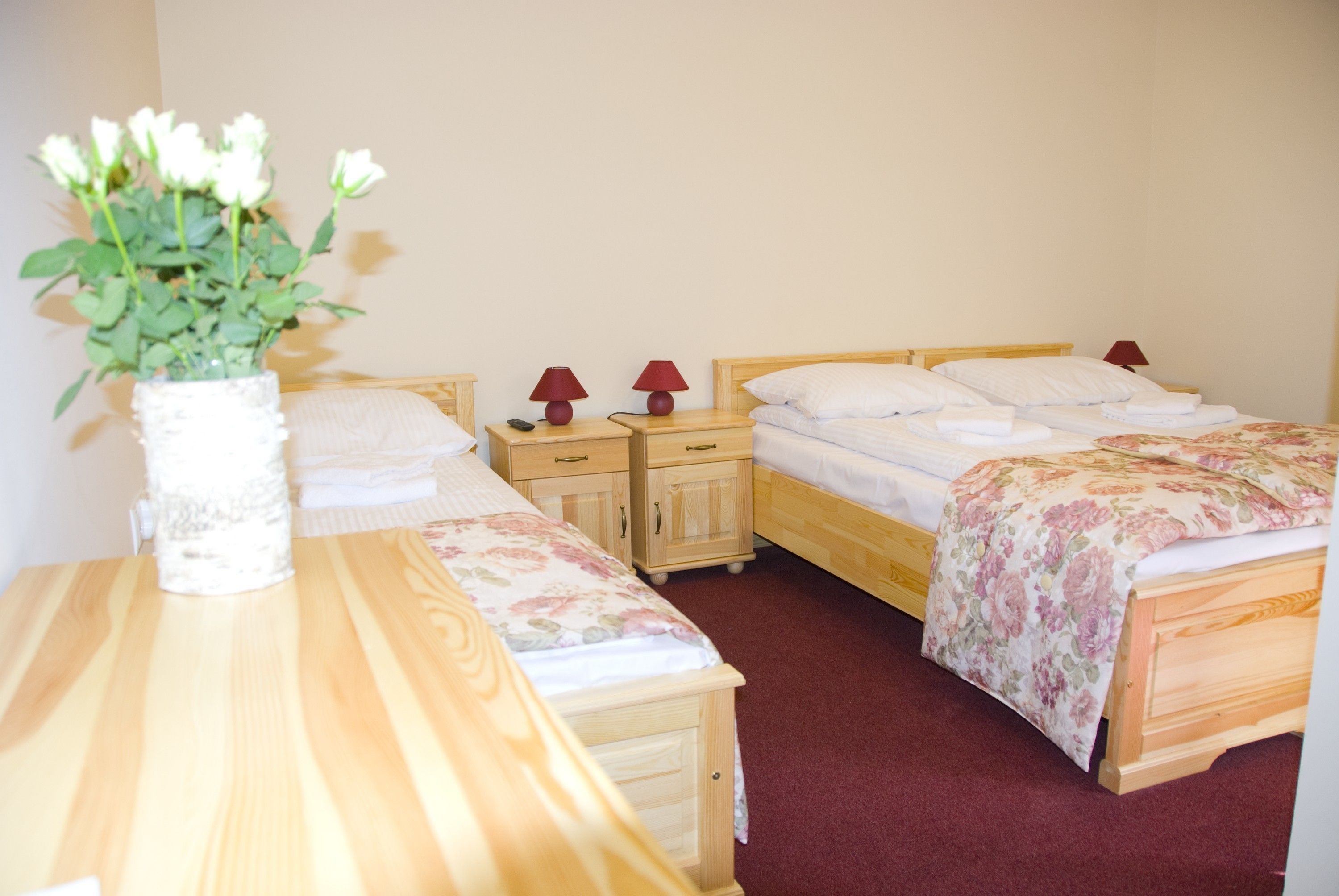 A hotel room with two single beds, a wooden nightstand with a vase of white flowers, and a wooden desk.