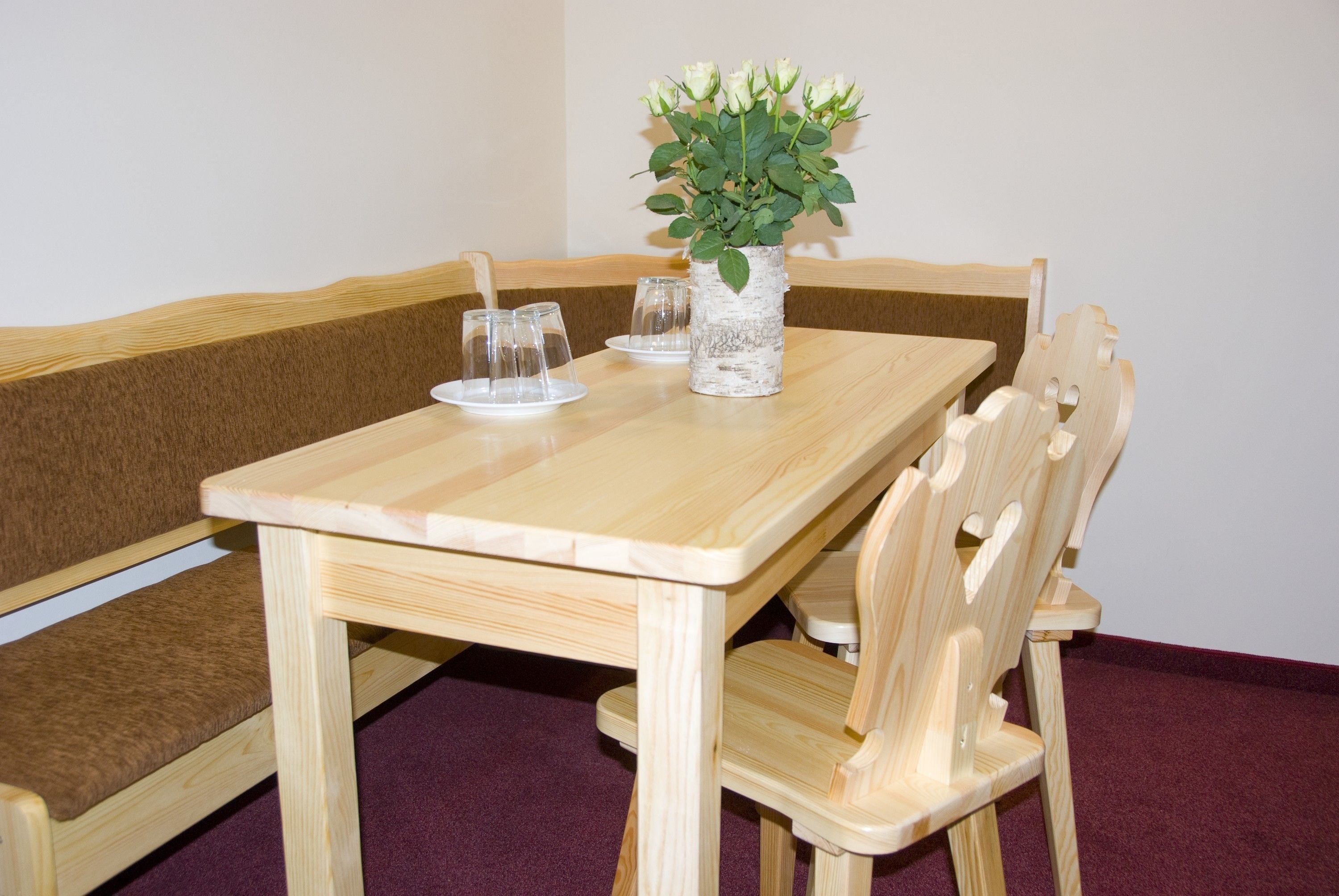 Una mesa de madera para comer con un florero de flores blancas, dos vasos y un plato.