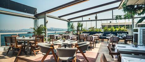 An outdoor dining area with tables and chairs, overlooking a body of water and cityscape.