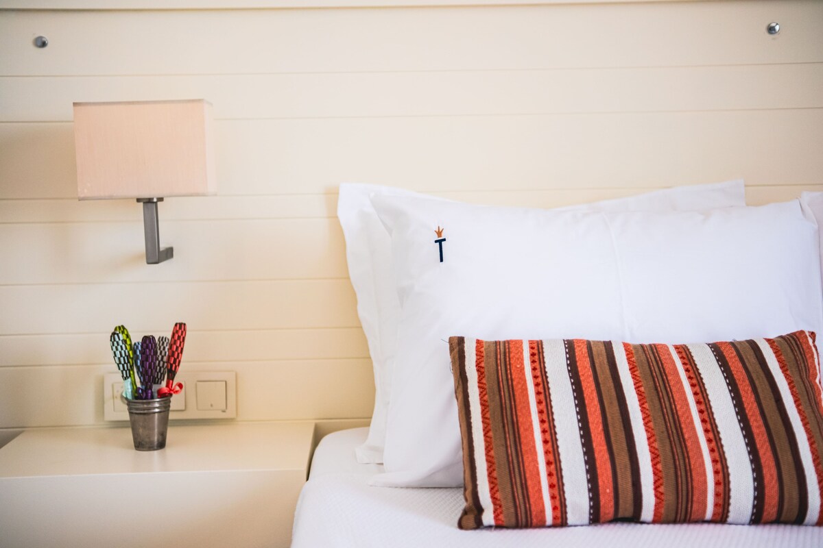 A hotel room with a bed, a bedside lamp, a nightstand with a cup of pens, and a striped pillow.