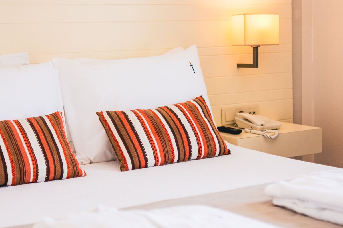Una habitación de hotel con una cama, dos almohadas a rayas, una mesita de noche con un teléfono y una lámpara de pared.
