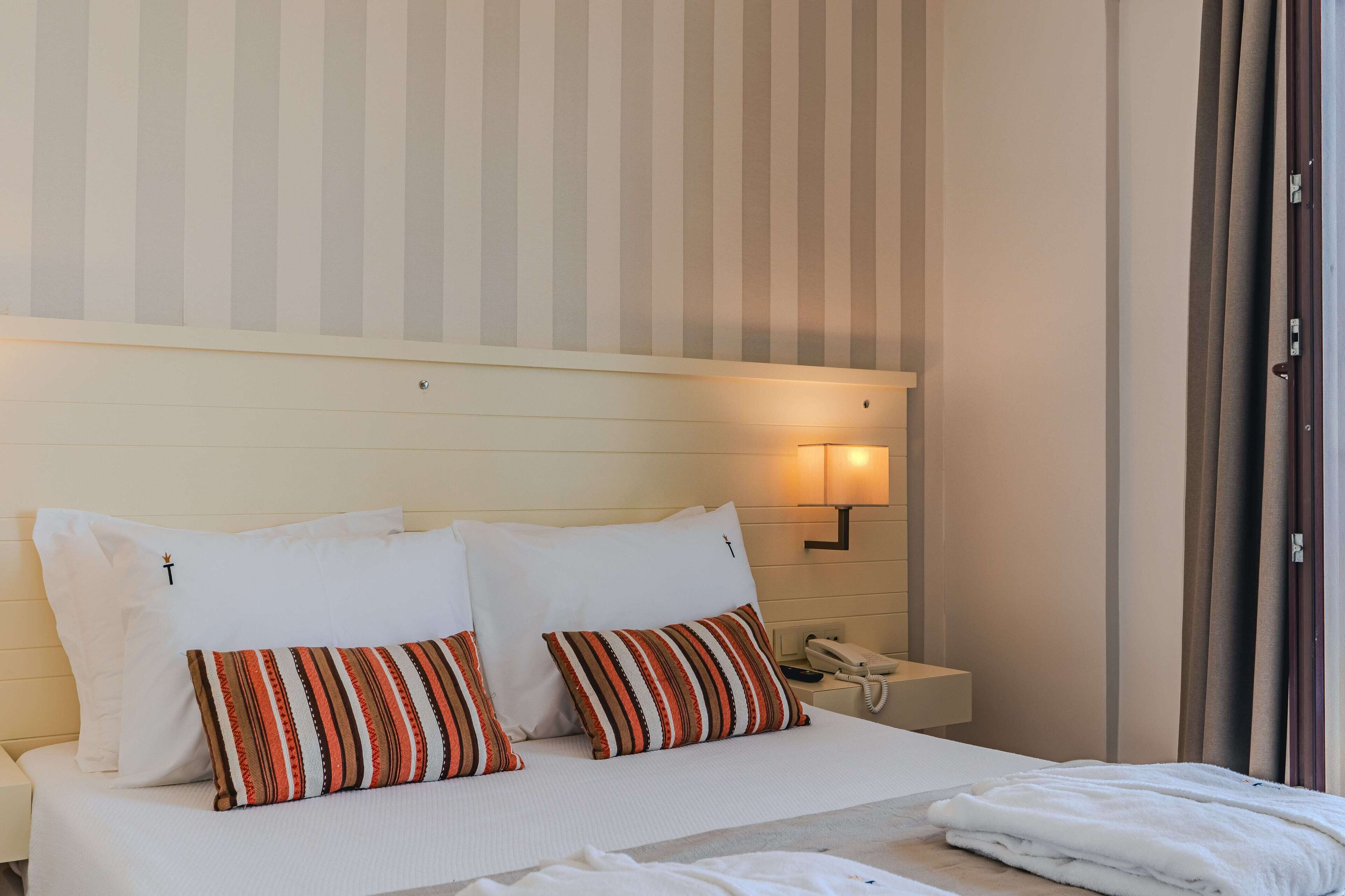 A hotel room with a bed, two pillows, a bedside table with a phone, and a wall-mounted light.